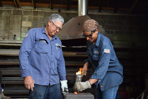 Two blacksmiths in navy work shirts form a piece of red-hot metal. One holds it in place while the other shapes it with a mallet.