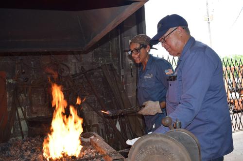 A young woman and a man, both wearing navy work shirts, hold metal tools to a fire.