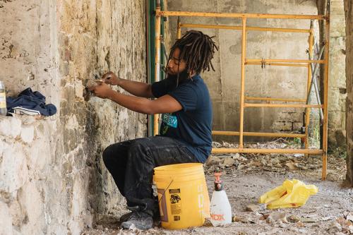 A man with long dreadlocks, seated, works on an old stone wall. 