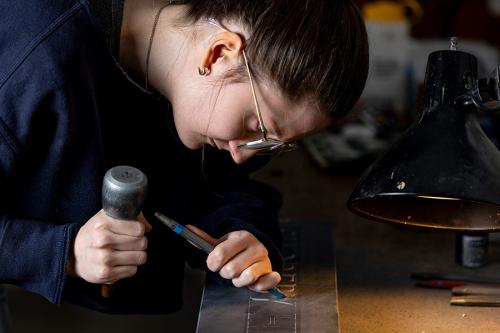 A young woman leans over a stone inscription in progress, holding a chisel in one hand and hammer in the other.