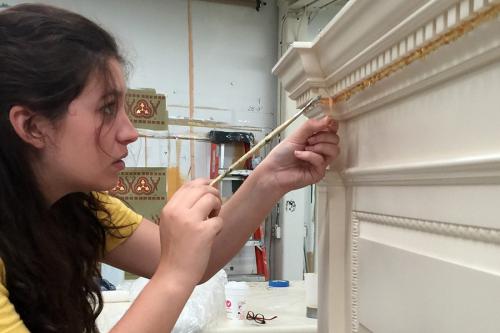 A young woman with a concentrated look on her face applies gold leaf to a white mantle with a paintbrush.