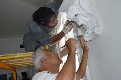 Two men work on a decorative plaster figure of a lion or other animal with open mouth, jutting out from a white wall. One man is standing on the ground, reaching up; the other is crouching down on a platform level with the decoration. 