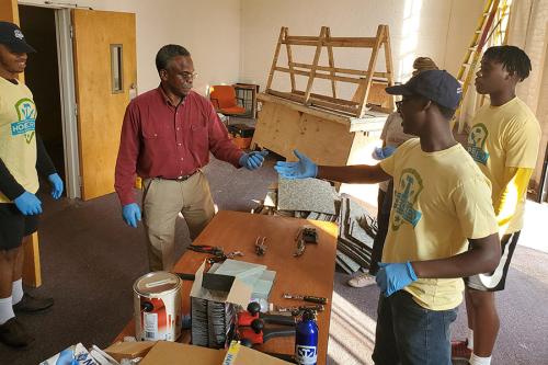 In a room surrounded by tools and wooden carpentry projects are three young men in matching yellow HOPE Crew T-shirts and an elder man in a red work shirt, all wearing blue latex gloves.