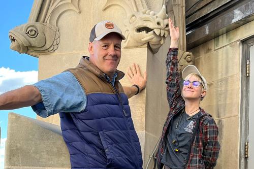 A man and a young woman pose next to decorative elements of a stone building, including heads of a fish and a dragon.