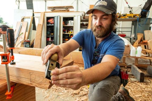 A young bearded man works in a carpentry workshop, using a metal tool on the corner of a piece of lumber.