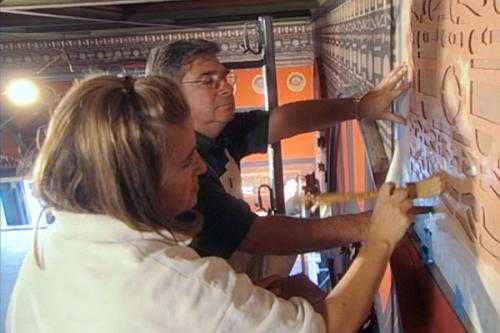 A man and a young woman use brushes on a stencil to embellish the moulding on a wall.