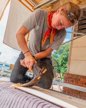 A young woman, safety glasses on her head and orange handkerchief around her neck, kneels as she uses a chisel on plank of wood.