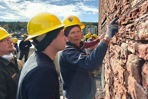 A man points to a detail in a red stone wall as a young person observes. Both wear yellow hard hats.