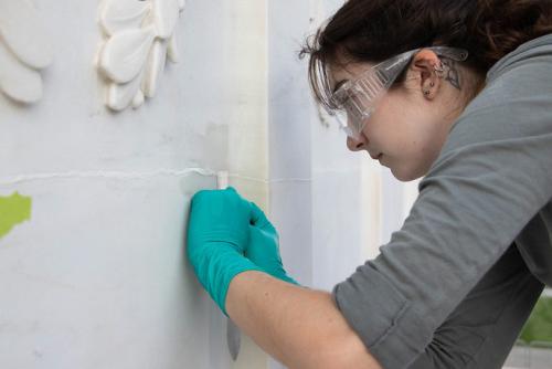 A young person wearing safety glasses and teal gloves uses a paintbrush on a horizontal crack across a white stone wall.