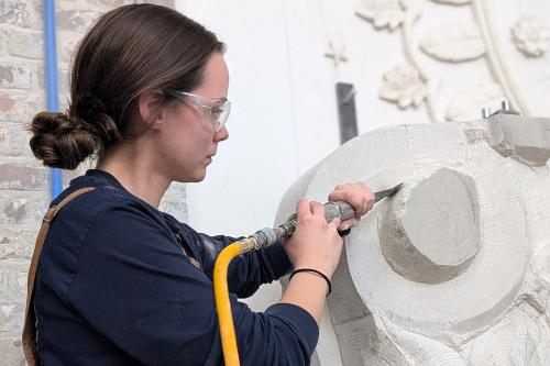 A young woman with long brown hair in a low bun and safety glasses uses a power tool to carve a large piece of white stone.