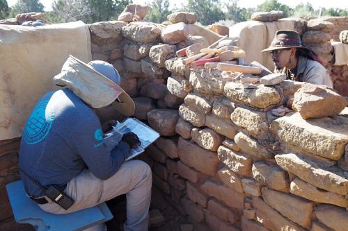 Two people work on either side of a low stone wall of a building with no roof.