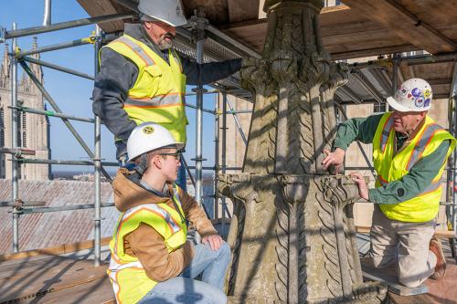 Three people in yellow safety vests and hard hats gather around a gray, conical pillar surrounded by scaffolding on the exterior of a building.