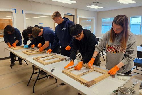 Six teens, all wearingg orange gloves, line a table as they work on rectangular wooden window frames.