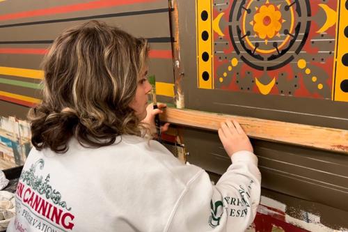 A young woman holds a long piece of wood along a brown wall to mark out horiztonal stripes. Above it is a stenciled pattern painted in yellow, red, and black, and to the side are stripes in red, black, yellow, and green. She holds a paintbrush in one hand