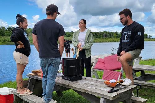 Four people gather around a big black pot on a portable stove on a picnic table at the side of a lake.