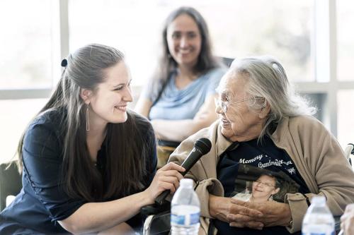 A younger woman interviews an Elder woman, holding a microphone up to her mouth. They both smile.
