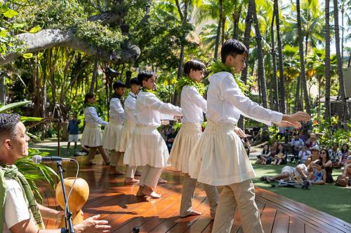 Boys in white perform hula on an outdoor stage in front of a crowd and palm trees.
