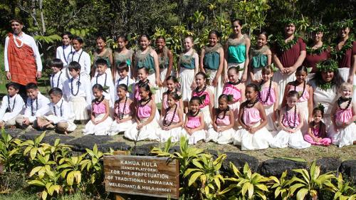Nearly forty children and a few adults pose in lines, wearing matching white, pink, turquoise, or dark red tops, beaded leis, and white bottoms. A wooden sign in front reads Kahua Hula (Dance Platform) for the perpetuation of traditional Hawaiian mele (ch