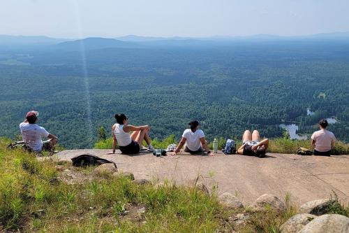 Five young people sit and lie along the paved edge of a summit, overlooking a vast forest landscape with mountains in the distance.