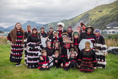 More than twenty adults and children pose in green grass, wearing matching black regalia with white and red fringe. Mountains and clouds loom in the background.