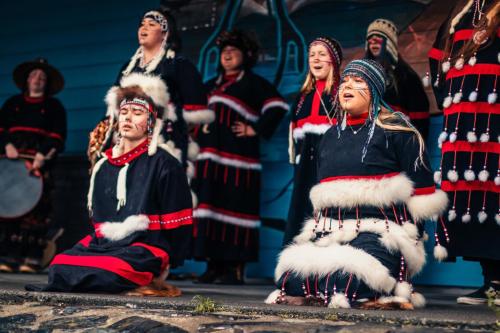Eight young people in matching black dress with red trim, white fur fringer, beaded caps, and lined face paint perform on stage.