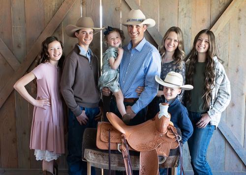 A family of two parents and five kids, including a toddler, pose in front of a brown leather horse saddle.