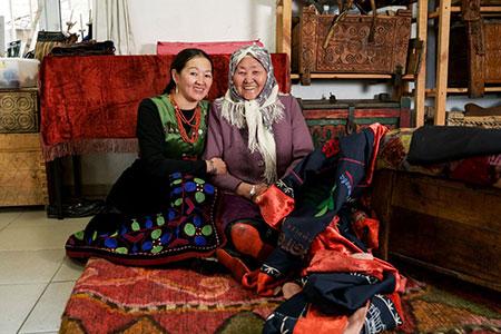 Two adults sit on the floor while holding each other and smiling at the camera. They are dressed in traditional outerwear from Kyrgyzstan.