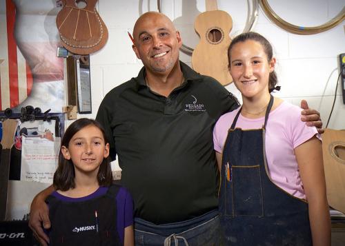 A father poses with his daughters on either side, each wearing aprons, in a workshop with parts of guitar-like musical instruments hanging on the walls.