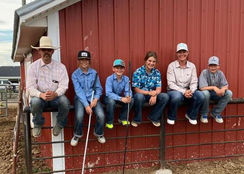 A family of two adults and four kids, all in jeans and Western-style shirts, pose sitting on a low metal fence along the side of a red barn.