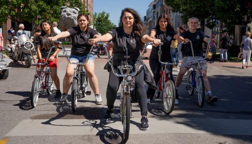 Five young people pose in a V-formation on lowrider bicycles with tall handlebars in a city street.