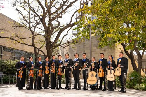 Thirteen young adults pose in black mariachi uniforms holding their instruments: violins, trumpets, and guitars.