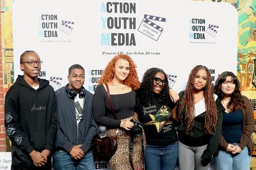 Sux young people pose in front of a white banner with repeated logos for Action Youth Media: Powered by Youth for 20 Years, and a graphic of a clapperboard with text: films with a focus.