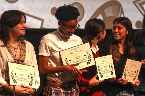 Four young people stand on stage, each holding up a framed certificate with a laurel design.