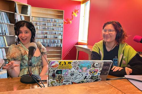 Two people sit in a radio studio with bright red walls and shelves of CDs and records. On the desk in front of them are two microphones and a laptop covered in stickers.