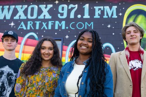 Four young people pose in front of a mural on the side of a building with the words WXOX 97.1 FM ArtxFM.COM in blue above a rainbow and over a starry black background.