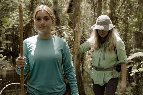 A young person hikes through the woods with a long walking stick.