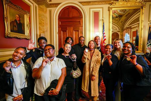 A group of eleven people pose, holding up medals around their necks. A portrait of George Washington hangs on the wall.