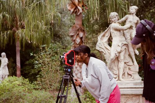 A young person hunches over to look through an analog camera on a tripod, standing in front of a marble sculpture of two figures in robes in a garden. Another person, partly out of frame, stands wearing headphones.