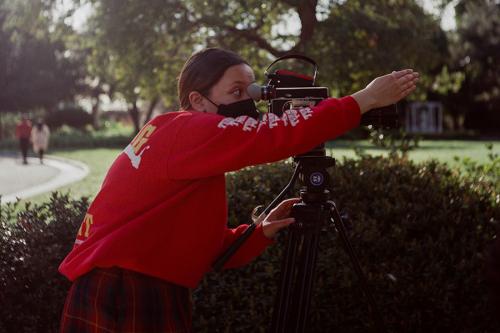 A young person looks through the viewfinder of a camera in a garden, pointing one hand straight ahead in the direction of the shot.