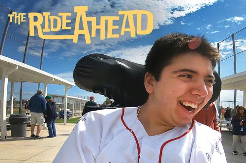 A young man smiles in his wheelchair, wearing a white baseball jersey with red trim and a signature on one shoulder. “The Ride Ahead” in yellow text is in the top left corner. 