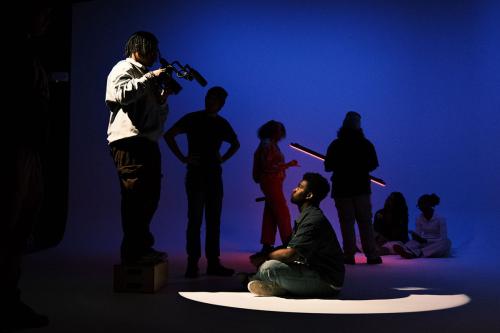 In a dark photo studio glowing with blue light, one person sits in a spotlight on the ground, looking up toward a person standing on a box, pointing a camera at them. Others sit and stand behind them.