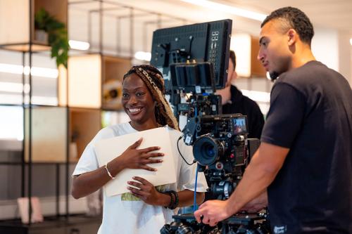 One person adjust a film camera with another stands behind the camera, smiling broadly.