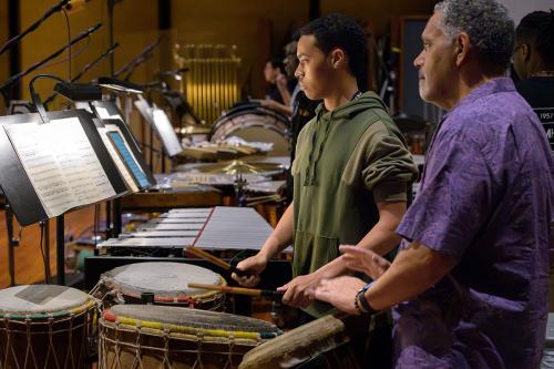 A young man in a green hoodie and an older man in a purple button-up shirt play barrel-shaped drums, reading from sheet music on a stand in front of them.
