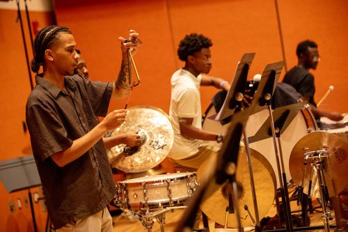 Four young men play percussion instruments in a music room: triangle, cymbals, and large drums.