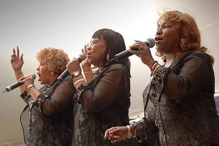 Three women in matching sheer black blouses sing on stage.