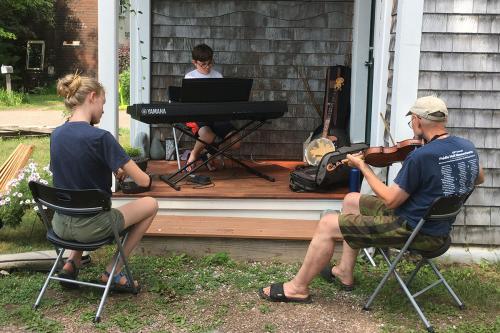 Sitting on and around a wooden porch, three musicians practice: a boy on keyboard, girl on fiddle, and elder man on fiddle.