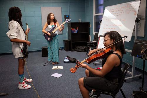 Three young people play music in a practice room with a whiteboard: electric guitar, electric bass, and violin.