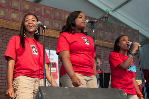 Three young women in matching red Stax Records T-shirts sing on stage under a festival tent.