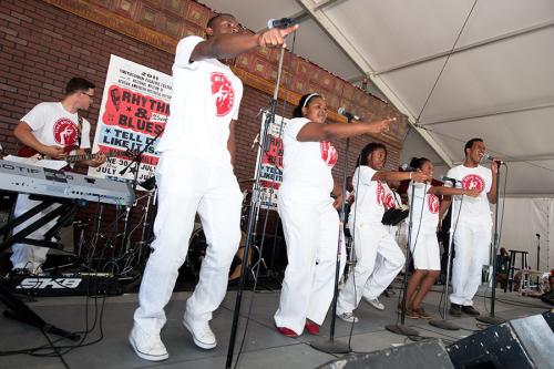 A band plays on stage under a festival tent, all wearing matching white shirts and pants. The five singers in front each point toward the audience.