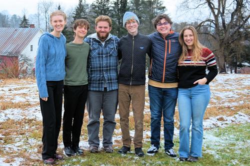 Six teens stand arm in arm on a grassy, snowy hill, smiling.